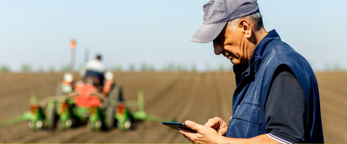 A farmer using a touchscreen tablet in a field, with another farmer operating heavy equipment in the